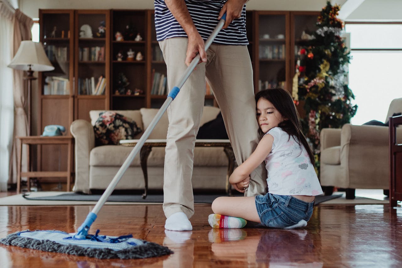 Father mopping the floor while daughter holds on, showing family bonding in a cozy living room.